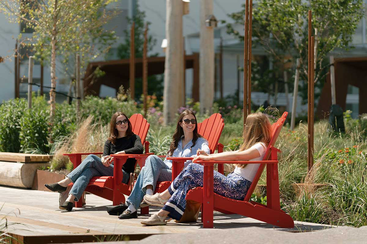 Visitors relaxing in bright red Adirondack chairs at a landscaped riverside plaza in New Westminster, BC, enjoying a slower pace during the fall season.